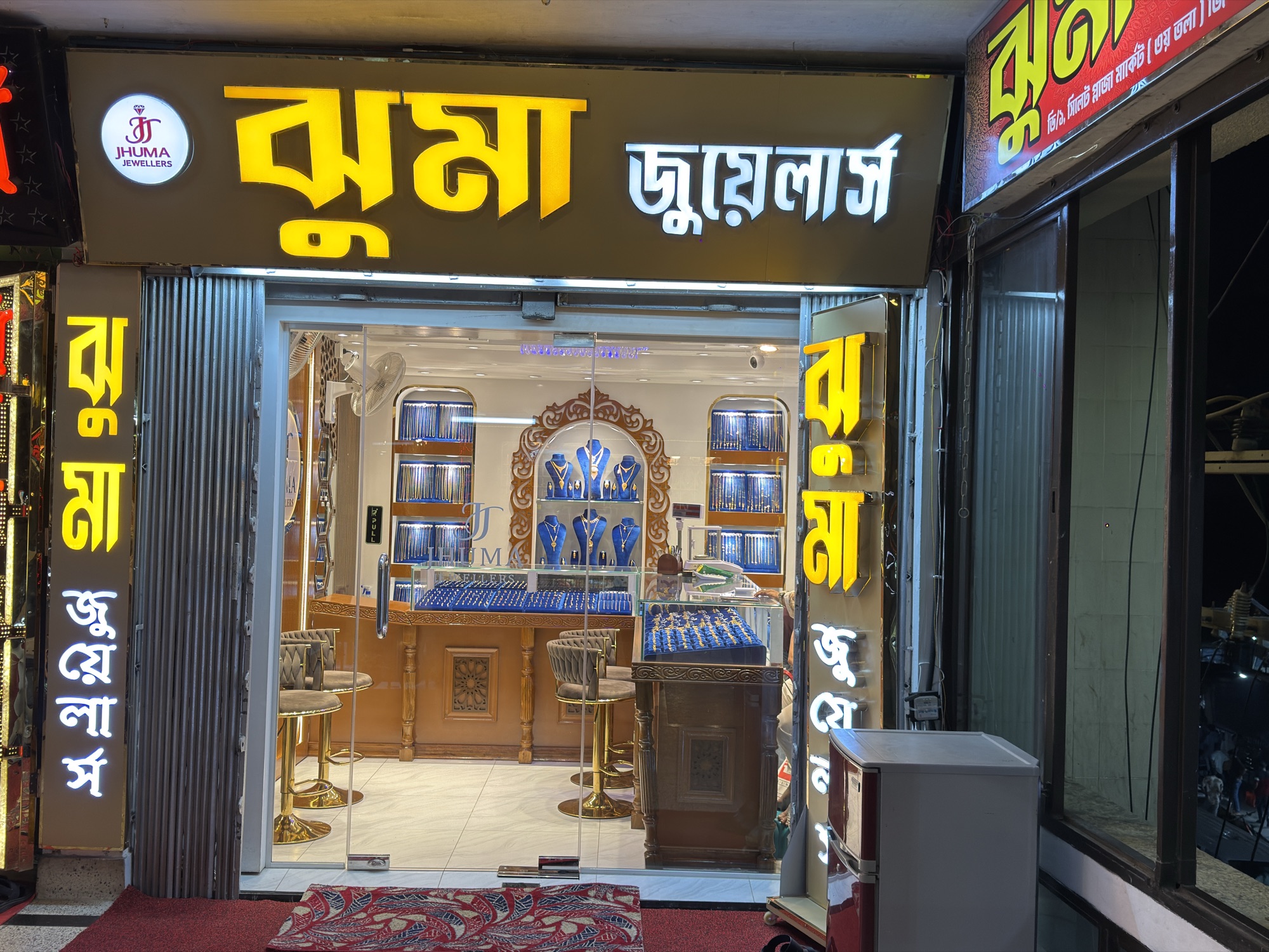 Night photograph of the Jhuma Jewellers storefront with blue neon and gold lettering.