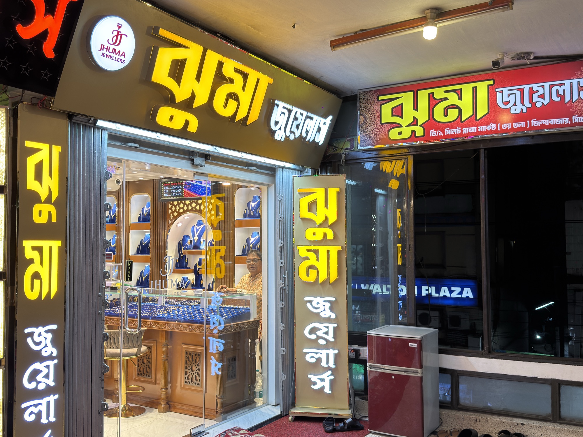 Wide night view of the Jhuma Jewellers glass front in Sylhet Plaza.